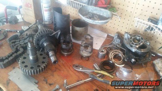 Assorted vehicle transmission and drivetrain parts including gears, a chain, bearings, and seals laid out on a wooden workbench
