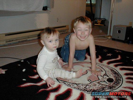 Two young boys on a rug with a moon and sun design. The older boy is shirtless and on his knees, smiling. The younger boy is wearing a long-sleeved shirt and pants
