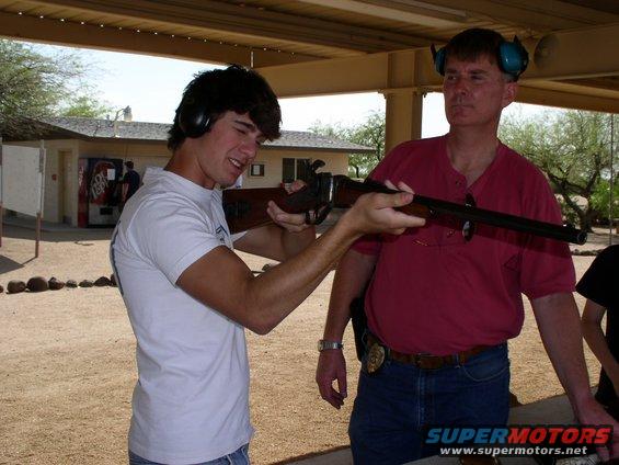 Young man aims rifle with instructor watching at outdoor shooting range