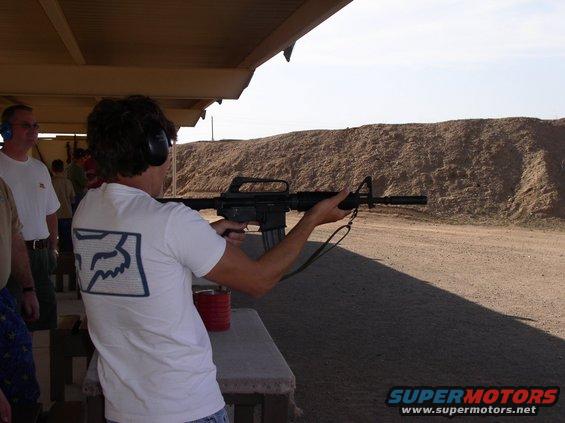 Man in white t-shirt with Fox logo aims rifle at outdoor shooting range