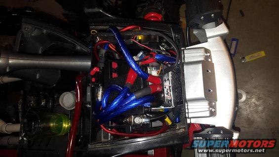 Vehicle battery compartment with blue and red wires, a battery, and a silver component. Some text is visible on the battery