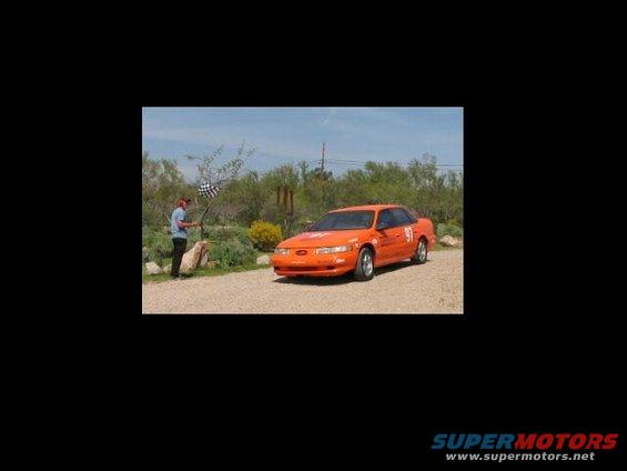 Orange 1993 Ford Taurus with racing numbers 91 on the door and fender, parked on gravel with a person holding a checkered flag