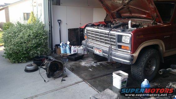 Red Ford Bronco with hood open, transmission removed, parked in driveway