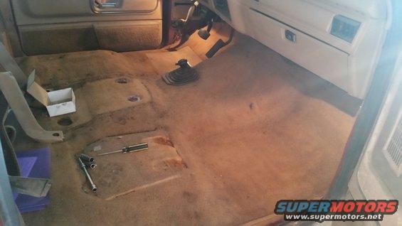Interior floor of a vehicle with carpet removed, showing transmission tunnel and some tools