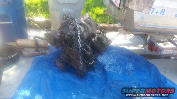 Engine assembly covered in chain, resting on a blue tarp. Part of a trailer and a trash can are visible
