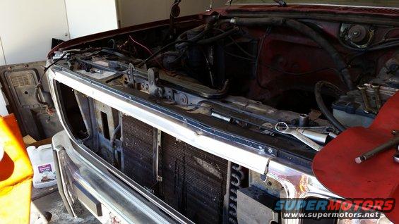 Red vehicle's engine bay with grille removed, showing radiator and engine components. Tools are visible