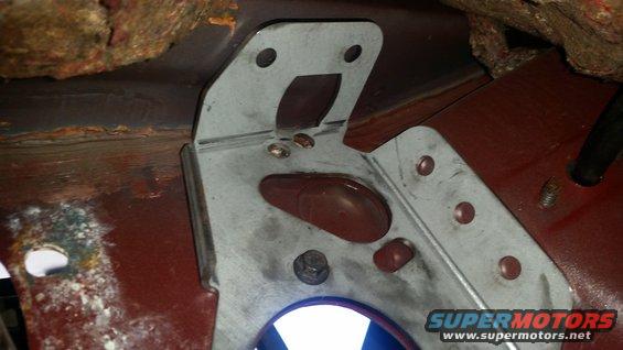 Metal bracket attached to a rusty red floor pan with visible bolts and a rubber grommet