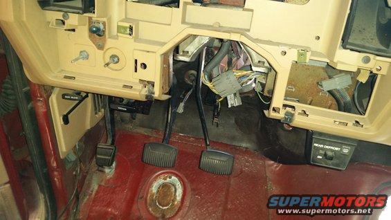 Vehicle interior showing dashboard, pedals, and wiring. Tan dashboard, red floor, and exposed wires are visible