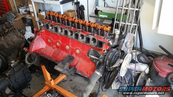 Red V8 engine block with orange valve covers on an engine stand, next to another engine. Garage setting