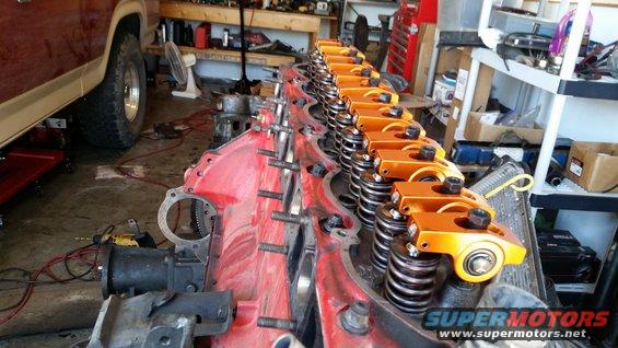 Red engine block with orange rocker arms and valve springs, partially visible vehicle in background