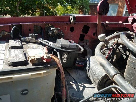 Close-up of a vehicle's battery and some engine parts. A black box labeled 'XGO' is visible near the battery terminals