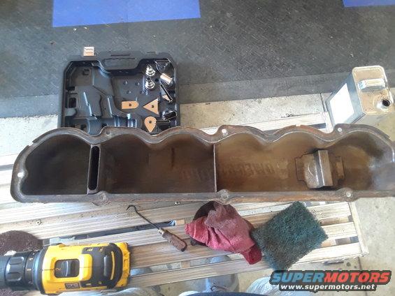 Engine valve cover with internal baffles, resting on a wooden pallet next to a drill and cleaning supplies