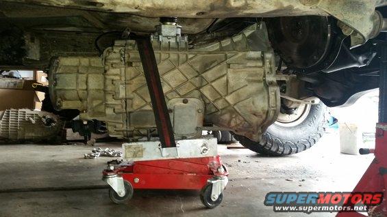 Transmission jack supporting a large transmission under a vehicle; tire and suspension visible