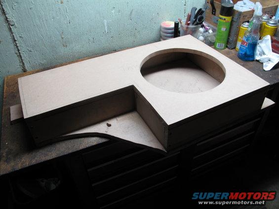 Unfinished wooden subwoofer enclosure with a large circular cutout, resting on a workbench