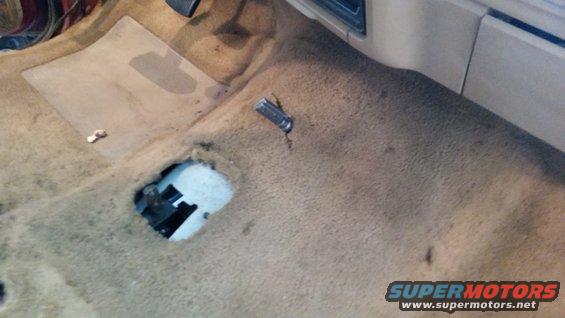 Interior floor of a vehicle with worn carpet, a hole revealing mechanical parts, and a metal object on the floor