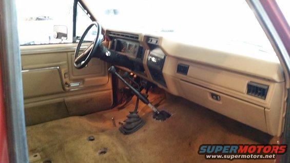 Interior shot of a red truck, showing the dashboard, steering wheel, and floor shifter. Tan interior panels and carpet are visible