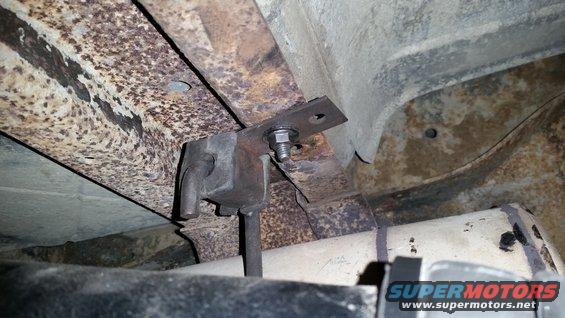 Underside of a vehicle showing rusty frame rails, a bracket with a bolt, and part of an exhaust pipe. Supermotors.net logo visible