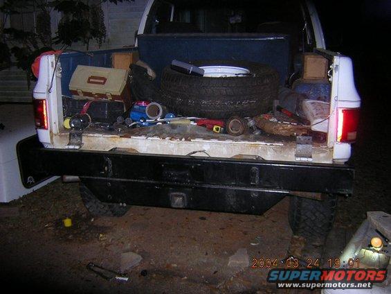 Rear view of a pickup truck bed loaded with tools, a spare tire, and other items. It has a black aftermarket bumper