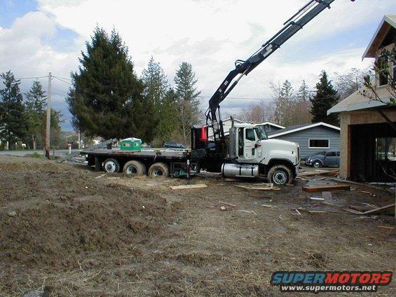 Flatbed truck with crane parked on muddy construction site next to unfinished house. Small car visible in background