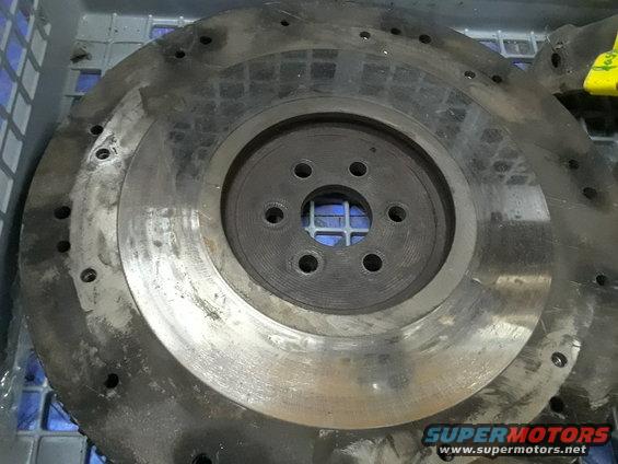 Metal flywheel with bolt holes and a central circular opening, resting in a blue plastic container