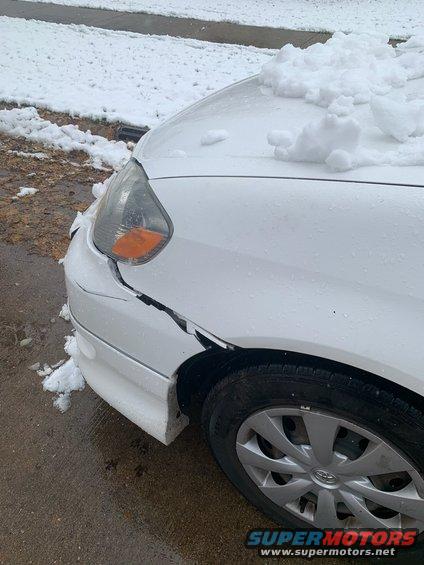White car front fender cracked and covered in snow, with a damaged headlight