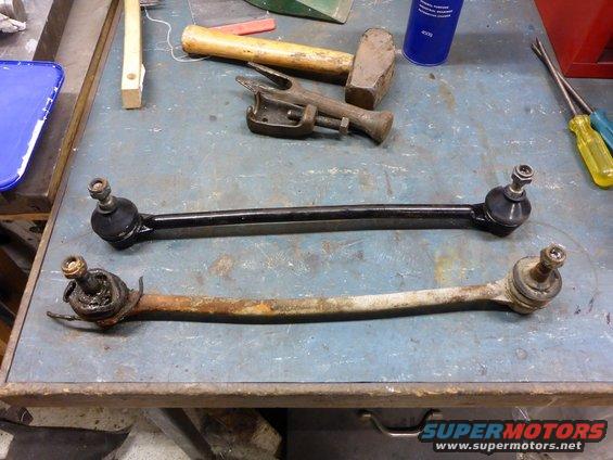 Two car suspension rods, one new and black, one old and rusty, lie on a workbench with tools