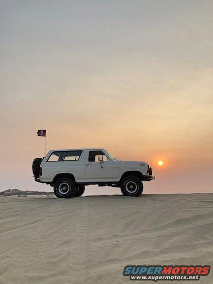 White 1980 Ford Bronco parked on a sand dune with a pink ribbon flag and a spare tire on the back