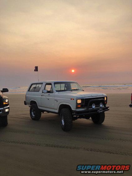 White Ford Bronco with off-road tires on a sandy beach, another vehicle partially visible to the left. Sunset in the background