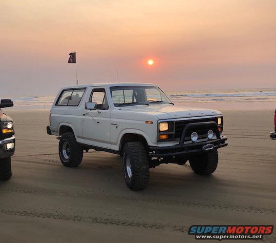 white Ford Bronco parked on a beach with the sun setting over the ocean