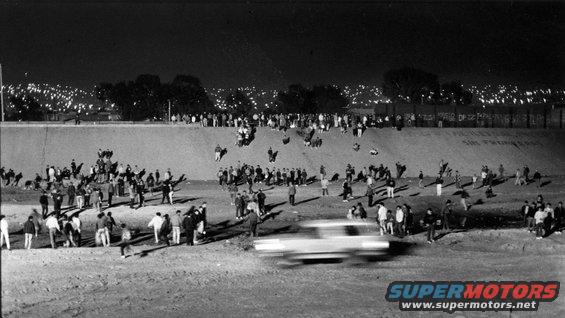 Blurry white car driving past a crowd at night with city lights in the background