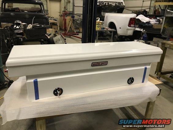 White Weather Guard truck bed toolbox on a table, with a black truck cab and a white truck body in the background