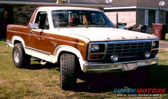 Brown and white Ford Bronco parked on grass in front of a house. It has a chrome grille and bumper