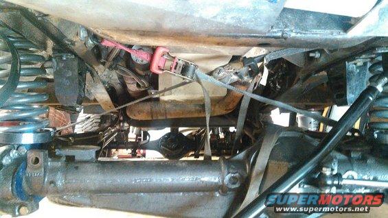 Underside view of vehicle suspension and drivetrain components, including axles and springs. Straps are securing some parts