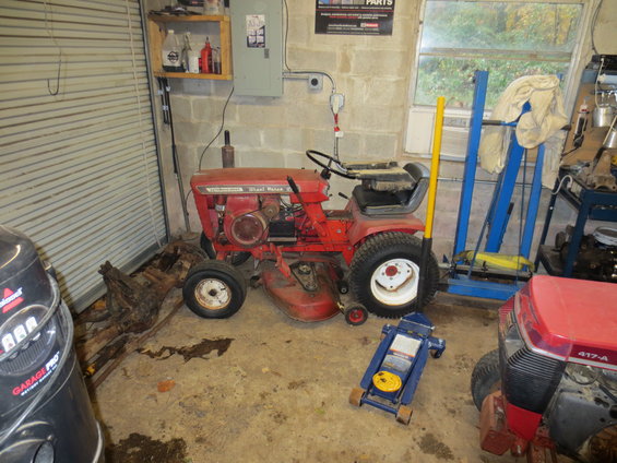Red riding mower with a deck, next to a blue floor jack. Another red mower is partially visible on the right
