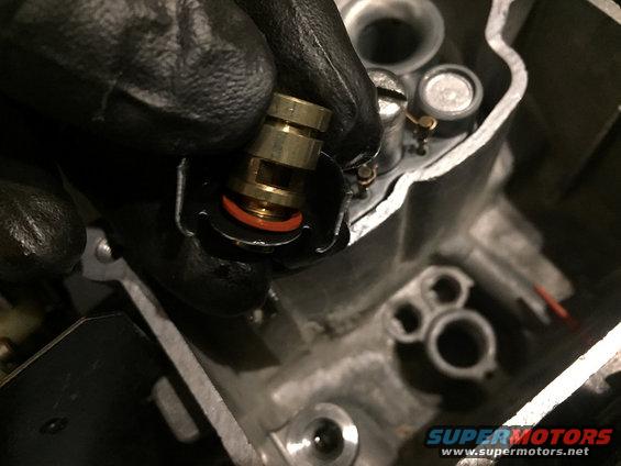 Gloved hand installing a brass component with an orange O-ring into a carburetor