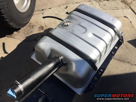 Aftermarket fuel tank with filler neck and straps, next to a tire