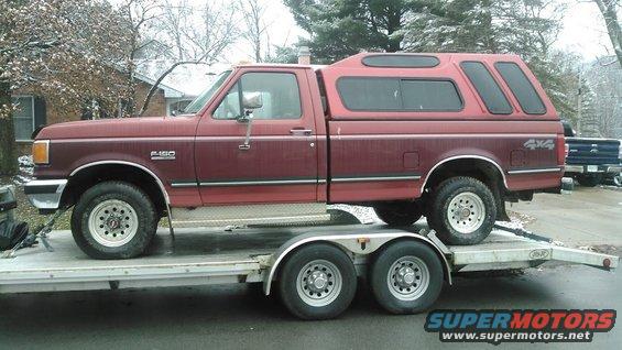 Maroon Ford F-150 pickup truck with a red camper shell, loaded onto a trailer
