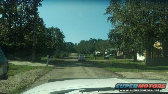 View down a rural road with two cars visible, surrounded by trees and houses
