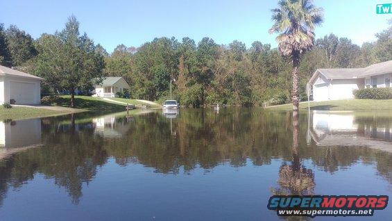 Truck submerged in floodwaters on a residential street, houses and trees in background