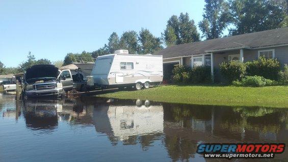 Front view of a dark-colored truck with hood up, partially submerged in floodwaters, with a trailer parked nearby
