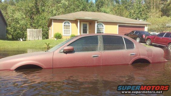 Maroon sedan submerged in floodwater in front of a house, with other vehicles visible in the background