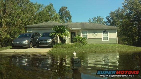 Two SUVs parked in front of a flooded house, one is a dark gray Honda Odyssey
