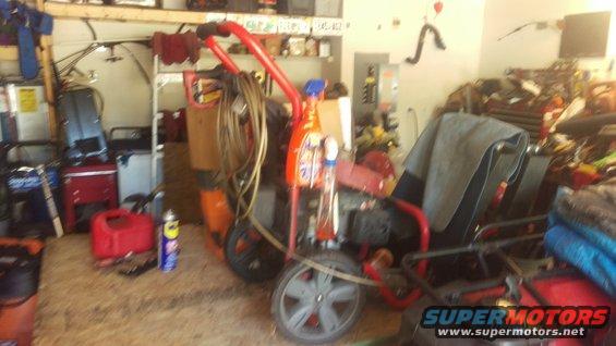 Red and orange pressure washer with coiled hose and cleaning bottles, parked in a cluttered garage