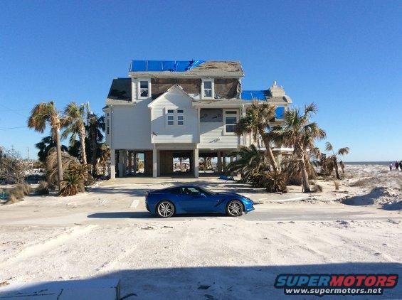 Blue sports car parked on sandy road in front of damaged house with blue tarps on roof