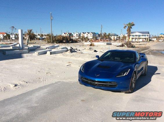 Blue sports car parked on sandy ground with damaged buildings in background