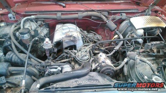 Engine bay with hoses, wires, and engine components visible. 'CAUTION FAN' text on a black shroud