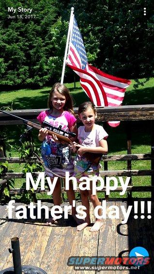 Two young girls stand on a wooden deck with an American flag behind them; one holds a rifle