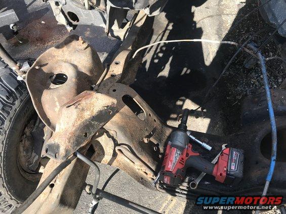 Rusty vehicle frame with a tire and impact wrench. The frame is heavily corroded and appears to be from a truck or SUV