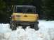 1990 Ford Bronco - Yellow Ford Bronco navigates a snowy trail, kicking up snow with its aggressive off-road tires.