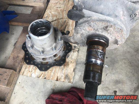 Disassembled vehicle axle and hub assembly on wooden surface. Supermotors.net logo visible